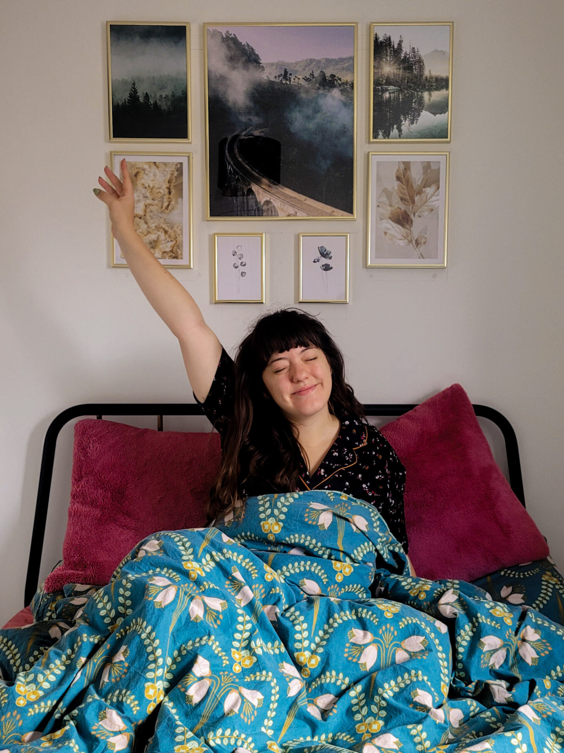 Jessica stretching her arms in the air, in a bed. A gallery wall of posters sits on the wall behind her head