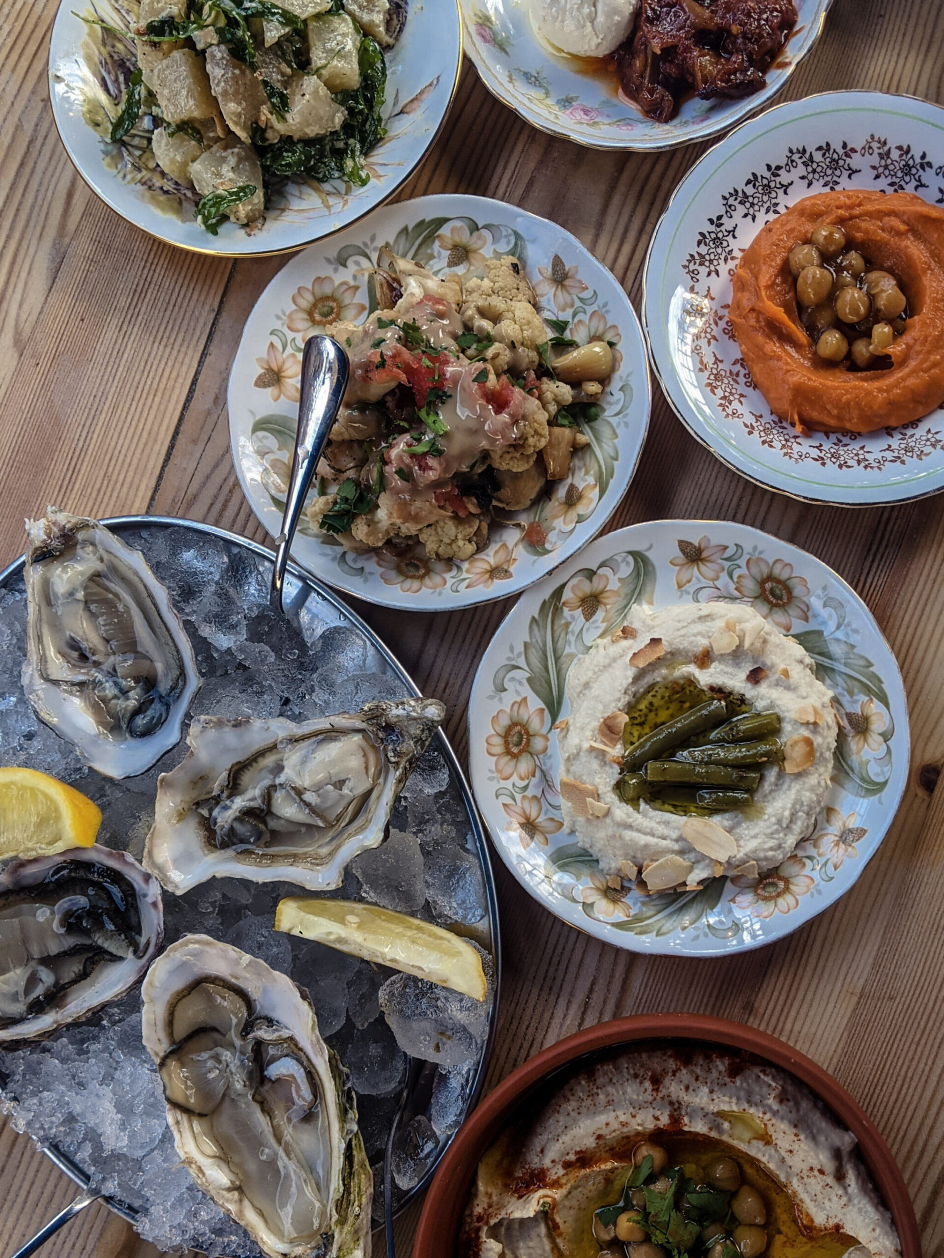 A plate of oysters, a bowl of hummus and chintz plates filled with vegetables, shown from a birds eye view